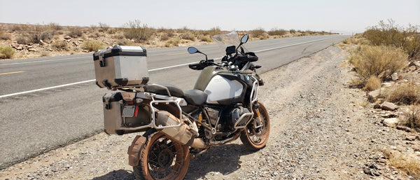 BMW R1250GS parked on desert highway shoulder in August heat, seat visible, empty road stretching into distance